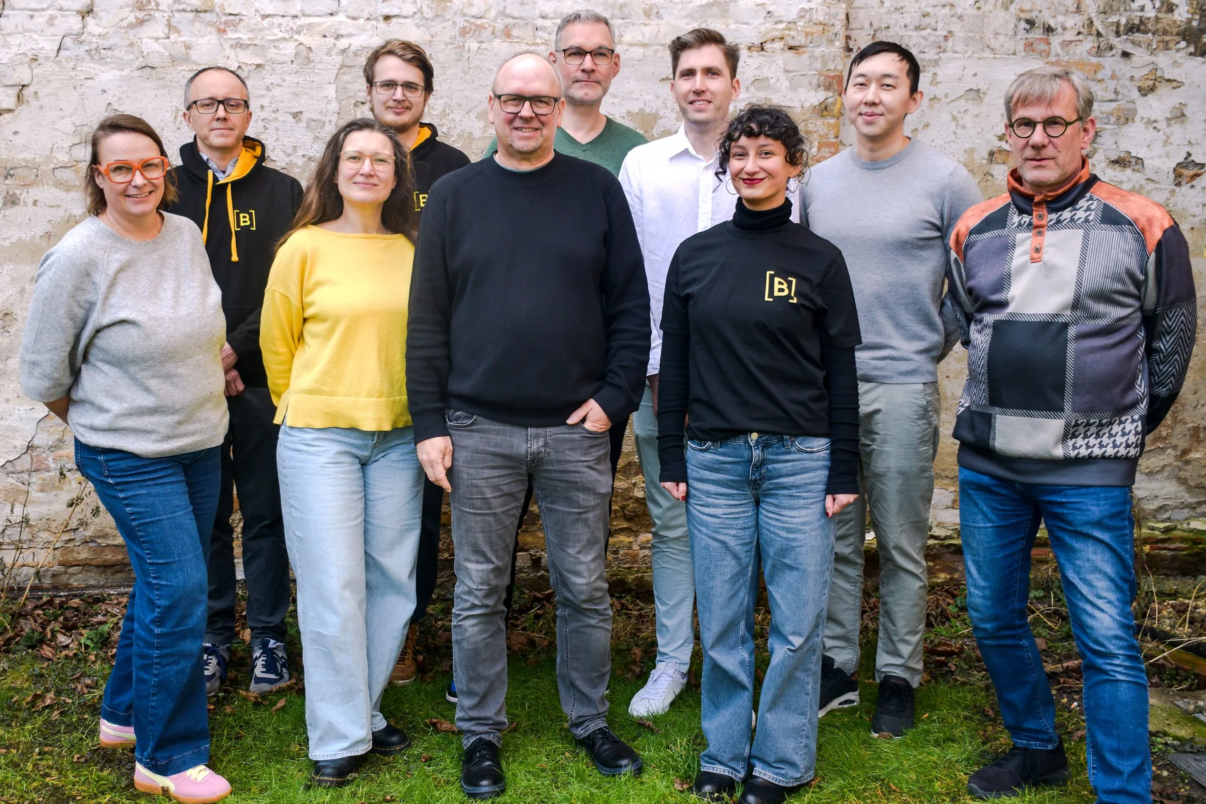 The ten members of the B-PROJECTS team standing together in front of a whitewashed brick wall